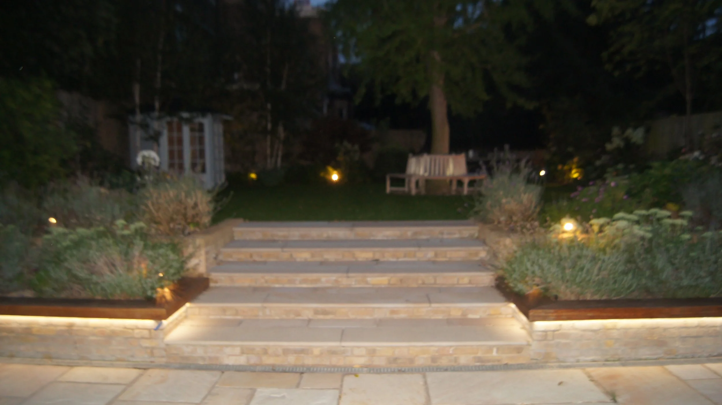 Nighttime backyard scene with stairs leading to a lawn, surrounded by plants and garden lights, with trees and a house in the background.