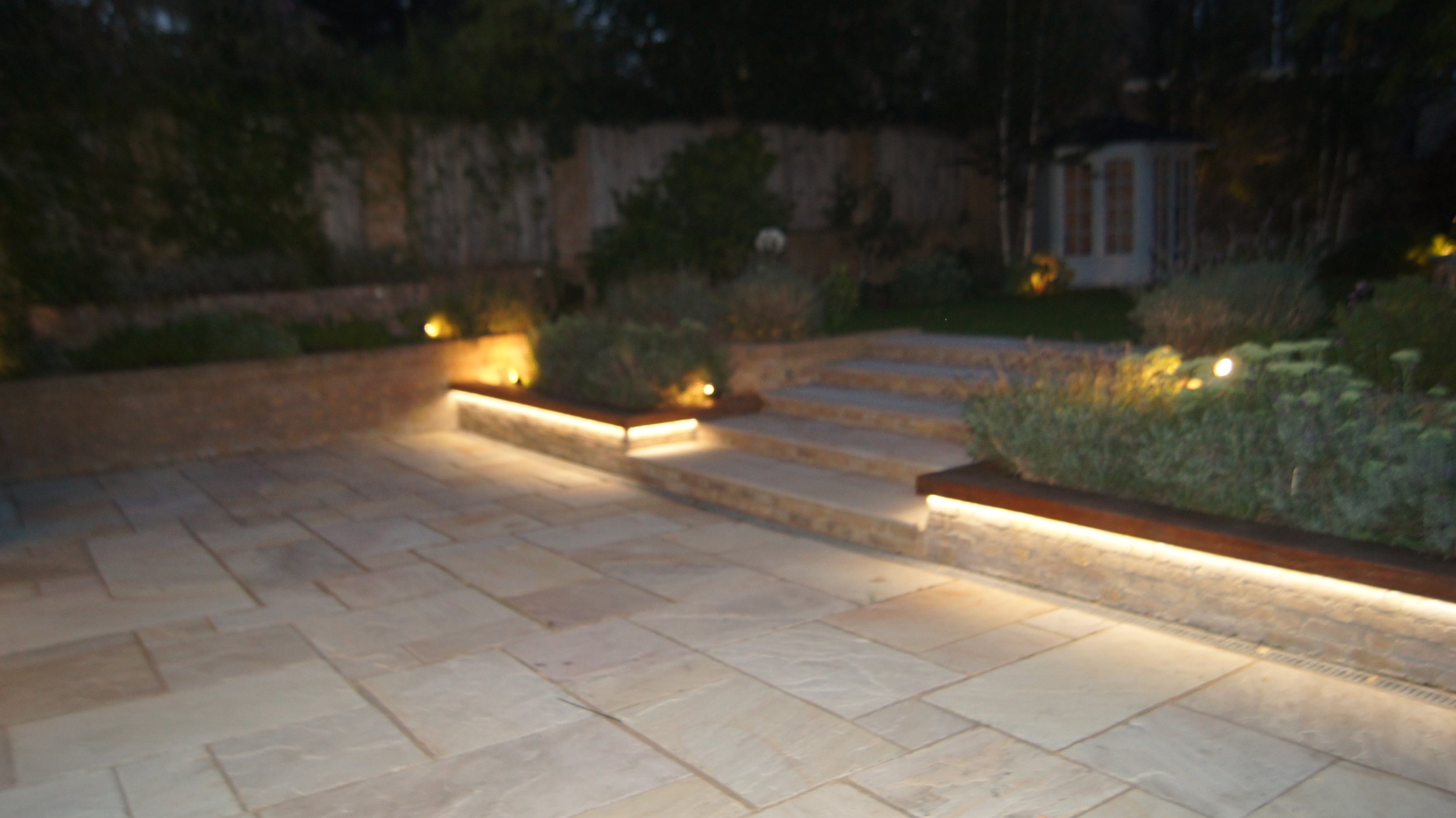 Nighttime view of an outdoor patio with stone paving, illuminated landscaped garden, minimal lighting, steps leading up to a grassy area, and a small structure in the background.