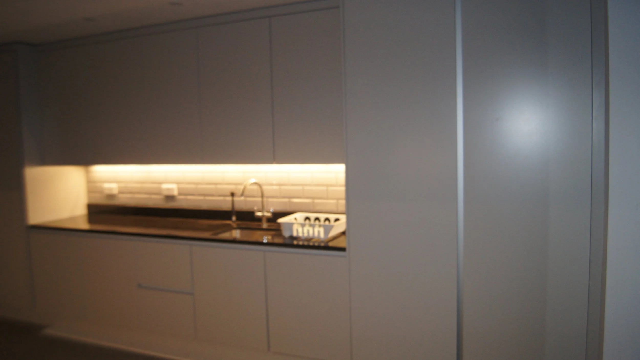 Modern kitchen with gray cabinetry, black countertop, sink, and a white dish rack, illuminated with under-cabinet lighting, and a tiled backsplash.