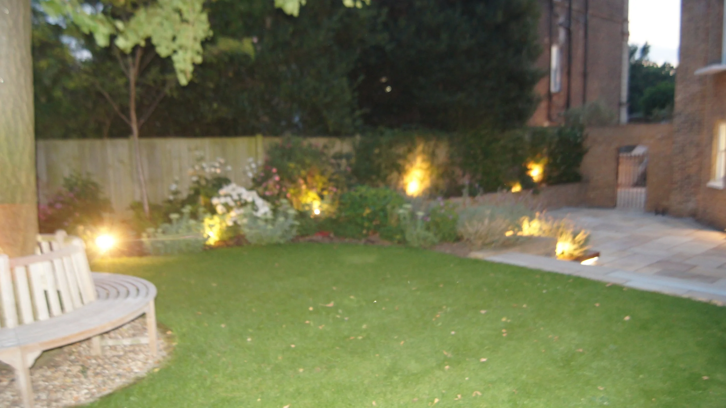 A backyard garden at dusk with a well-maintained lawn, flower beds, and exterior lighting illuminating the garden and brick buildings in the background.