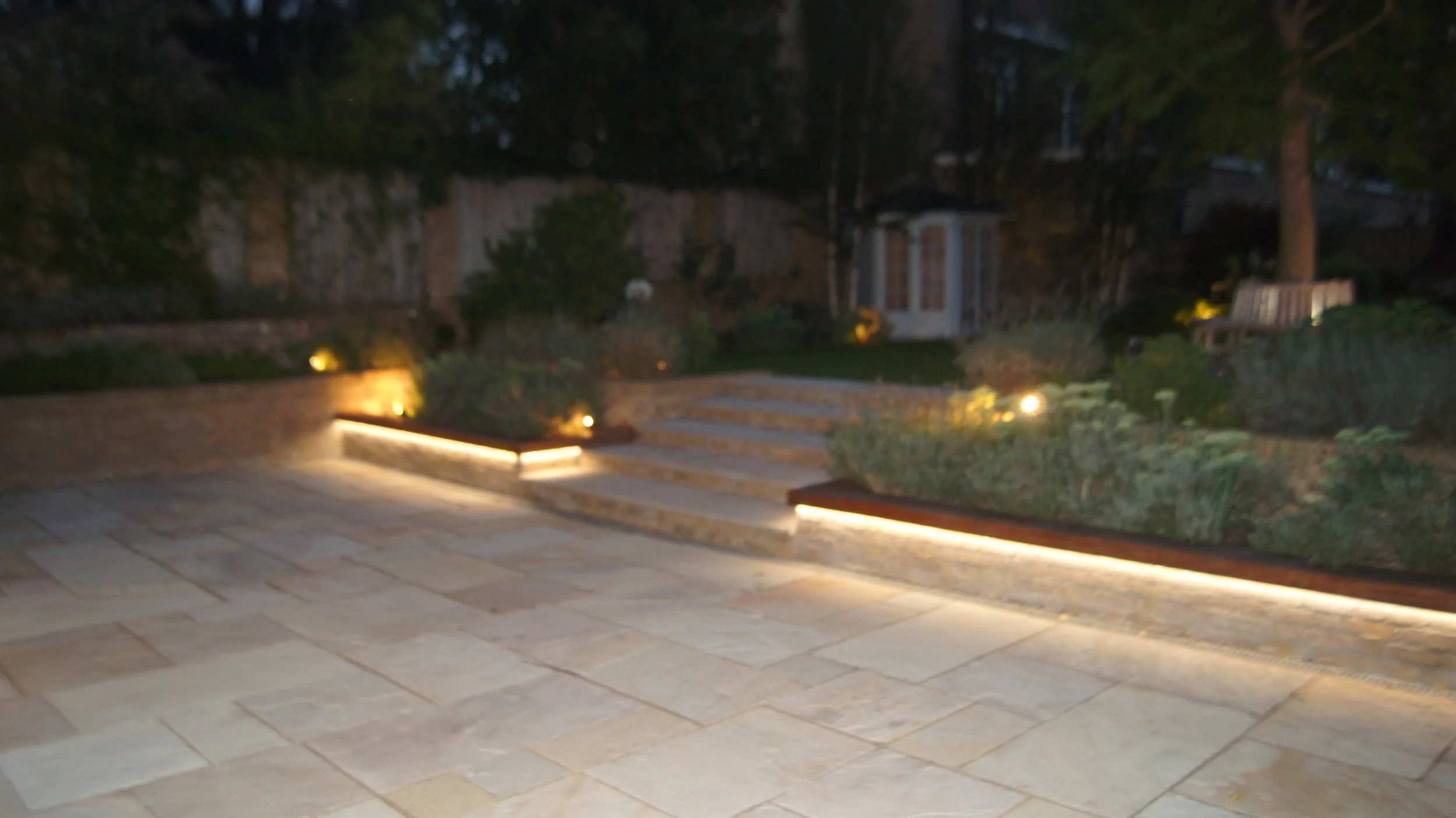 A backyard patio at night with stone paving, illuminated built-in lighting along the edges, steps leading to a garden with bushes, trees, and a small shed or playhouse in the background.
