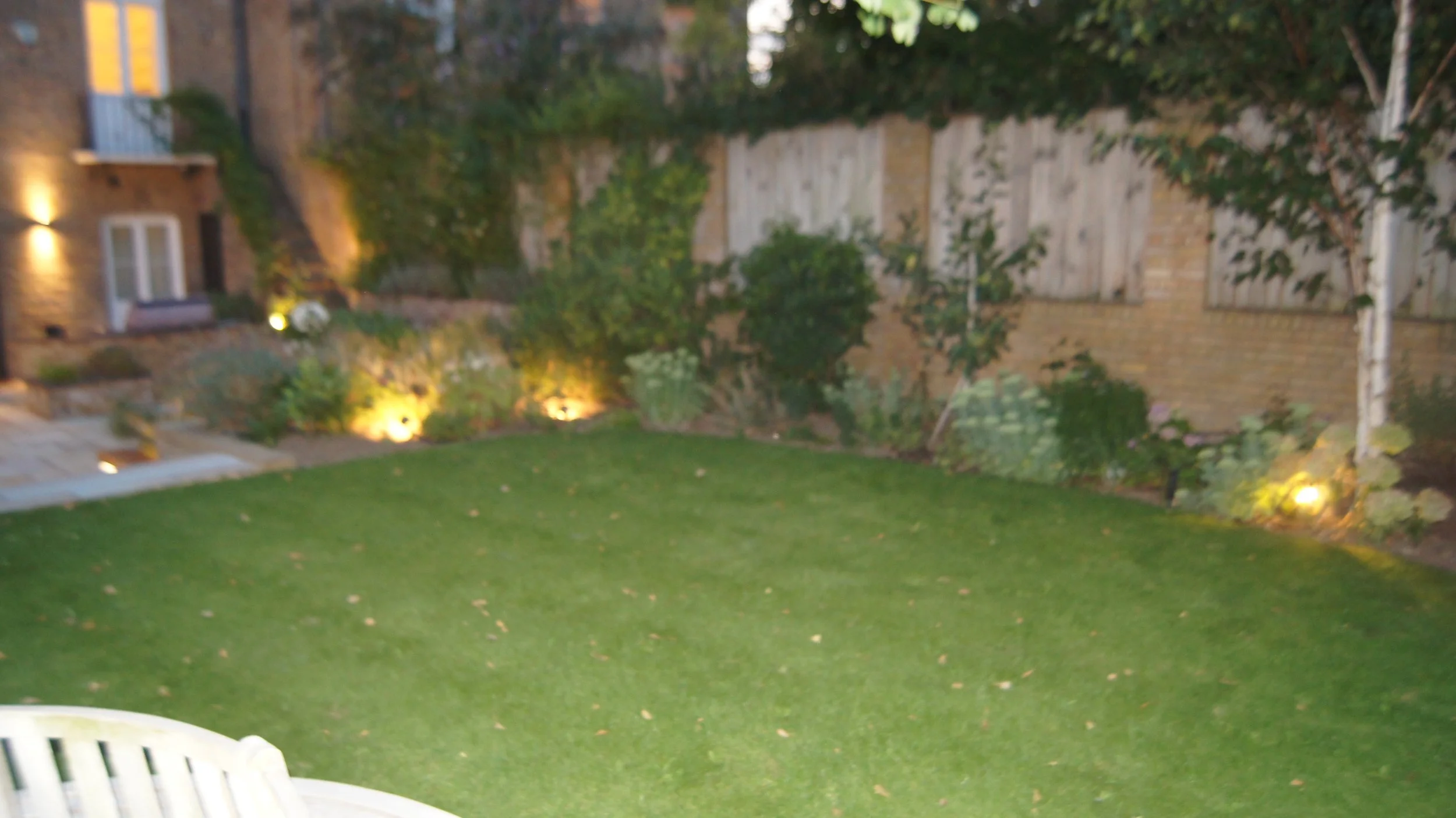 Blurred image of a backyard with a green lawn, garden beds, and trees illuminated by outdoor lights at dusk or evening.