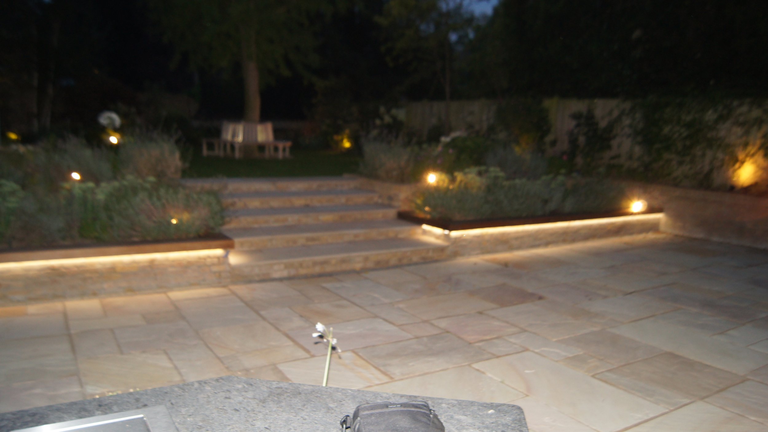 Nighttime outdoor patio with stone steps leading to a garden, illuminated by low-voltage landscape lighting. There are trees, bushes, and a wooden bench in the background.