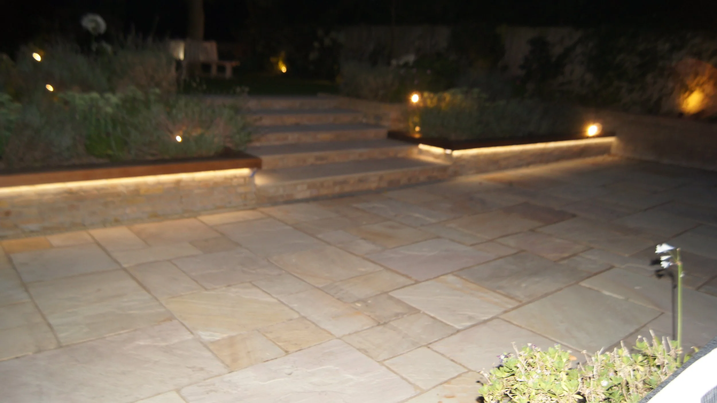 A stone patio with steps leading up to a garden area at night, illuminated by outdoor lighting.