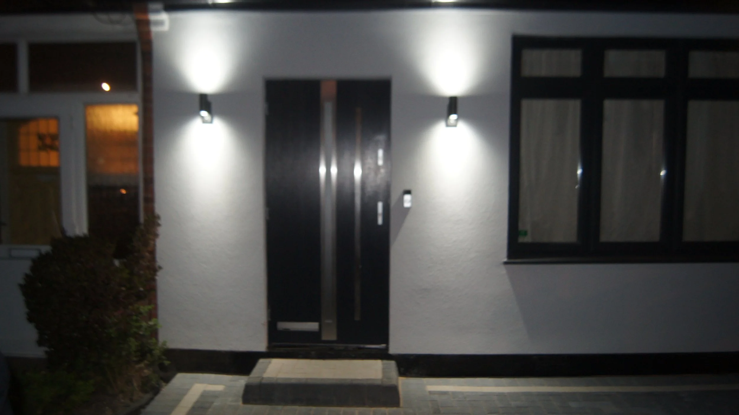 Nighttime exterior view of a house with a central black door, two wall-mounted lights illuminating the entrance, and a large window to the right of the door