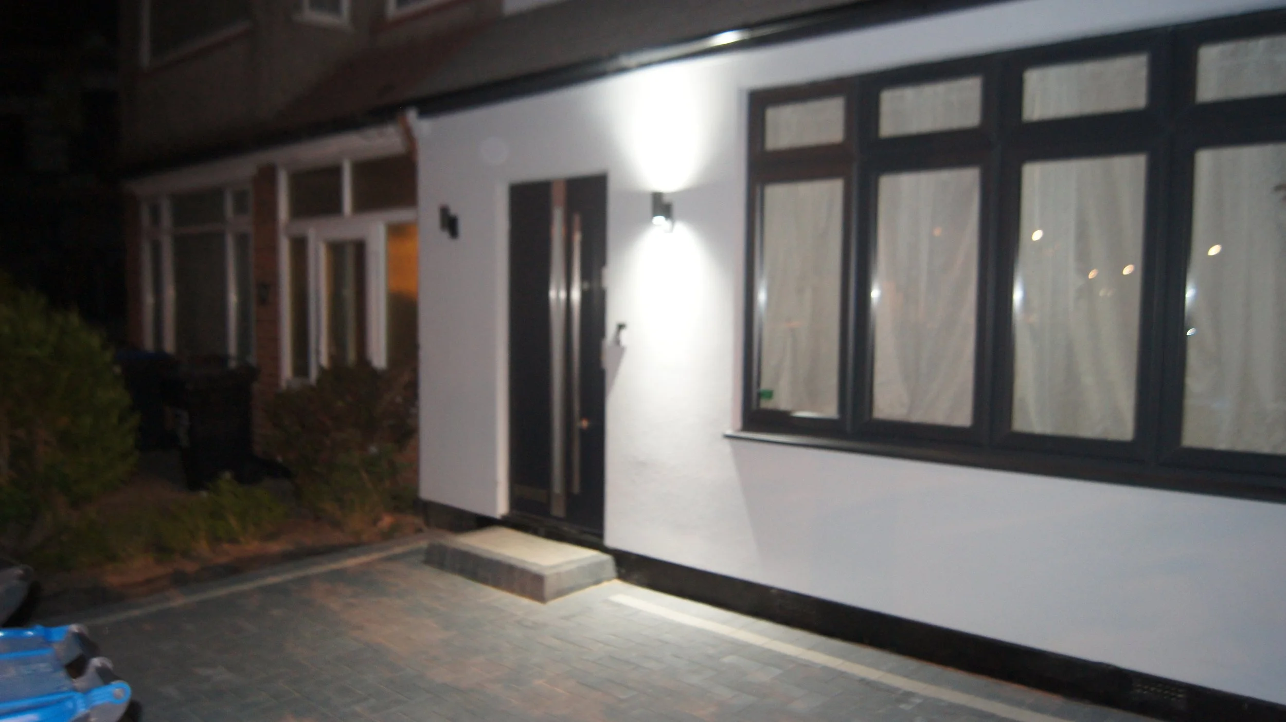 Nighttime exterior view of a modern house entrance with a black front door, a wall-mounted light above it, and a large window with curtains, showing a paved pathway and some shrubbery.
