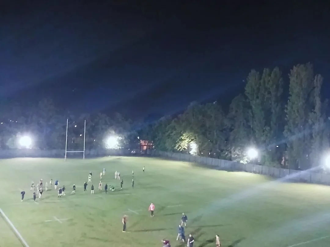 Night time view of a football field with players and spectators, surrounded by trees and lit by bright floodlights.