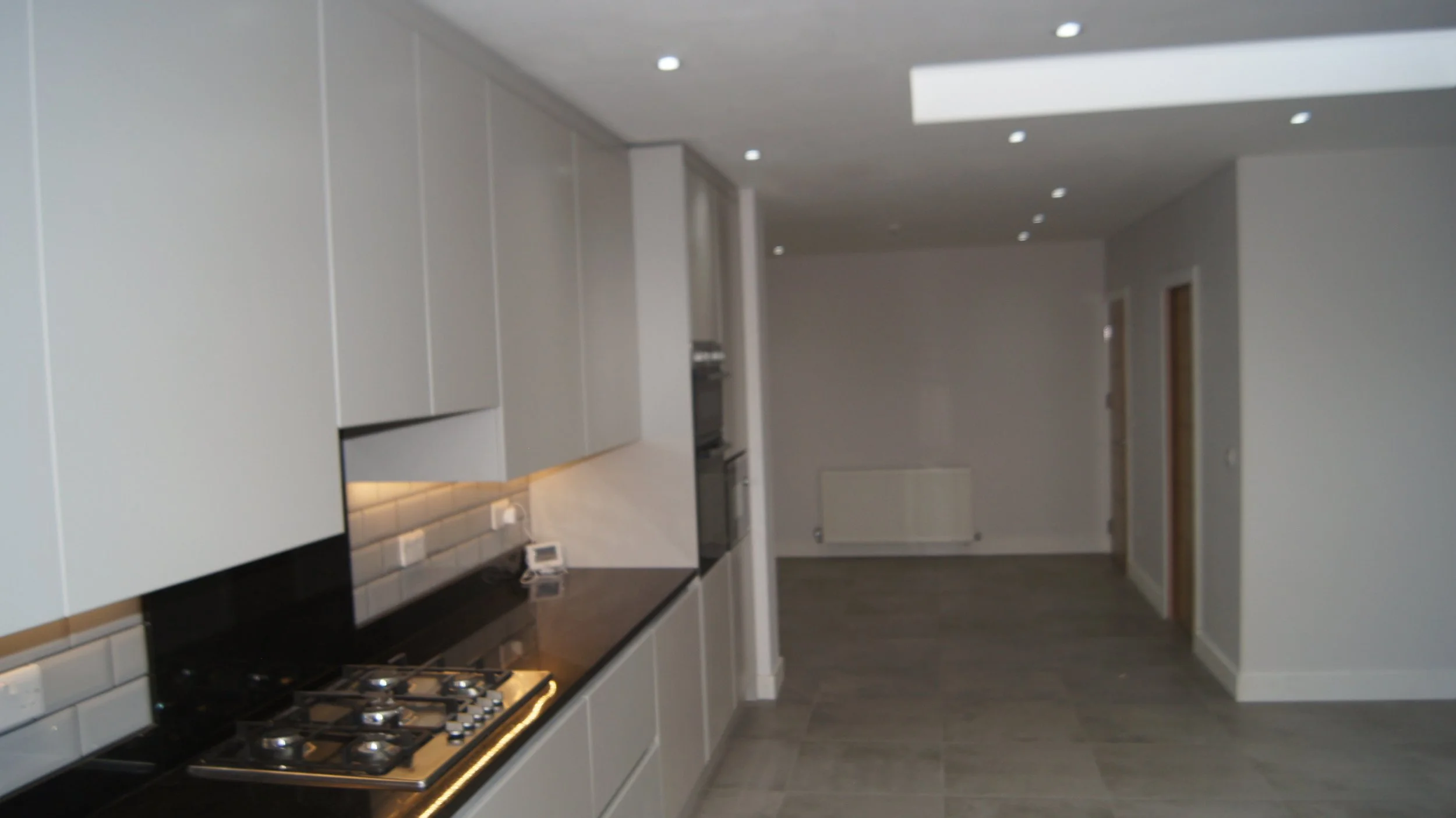 Empty modern kitchen with light gray cabinets, black countertop, and built-in oven, with an open living space beyond.