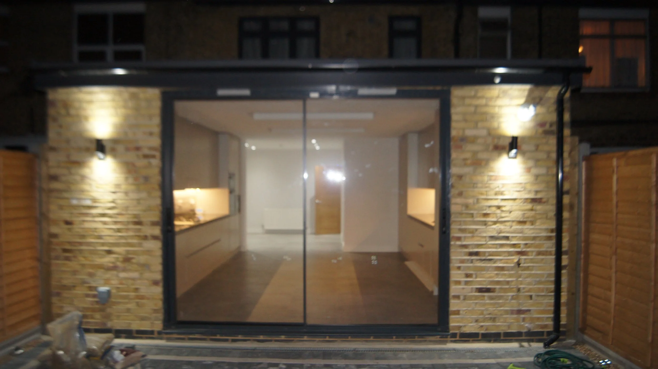 Night view of a modern building with a glass door and brick exterior, illuminated by outdoor wall lights, and a wooden fence on either side.