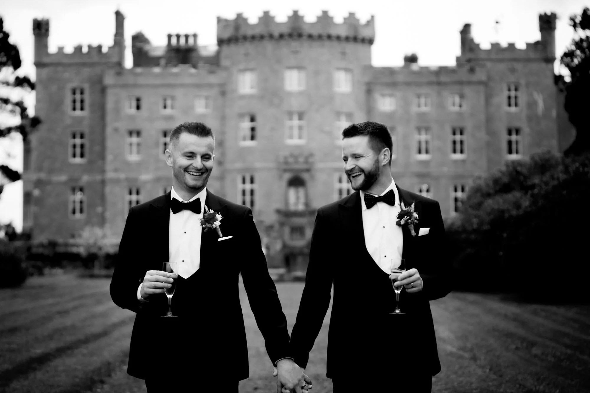 A black and white wedding photograph of two grooms walking away from their Irish castle wedding