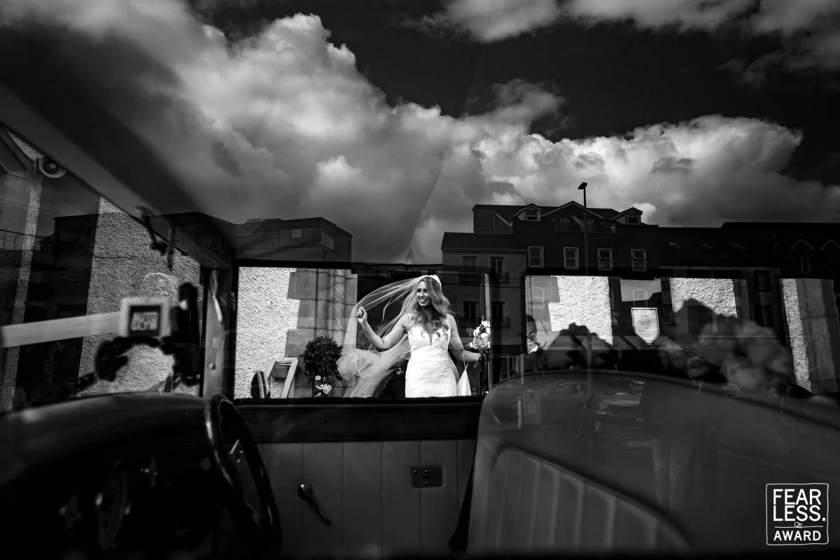 A creative wedding photographer uses the reflections of the bridal car to frame the bride leaving her Dublin wedding ceremony