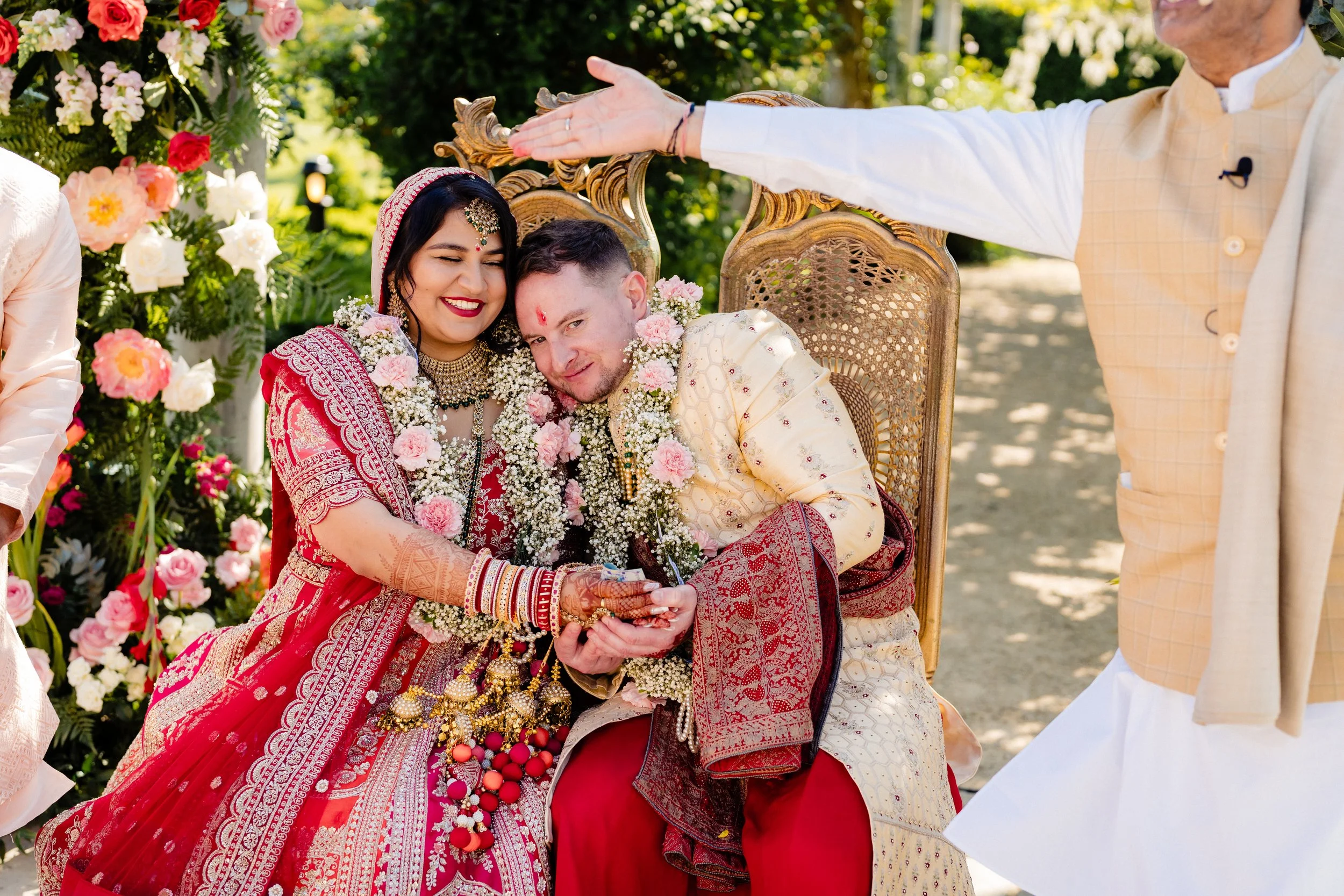Indian bride and groom exchanging rings during outdoor wedding ceremony, surrounded by floral decorations, with a man extending his hand from the right.