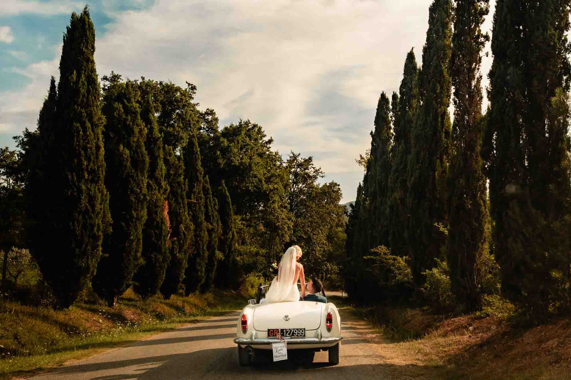 A bride sits on her convertible vintage car cruising through the dusty streets of Tuscany in this destination wedding photography by Annie Kheffache