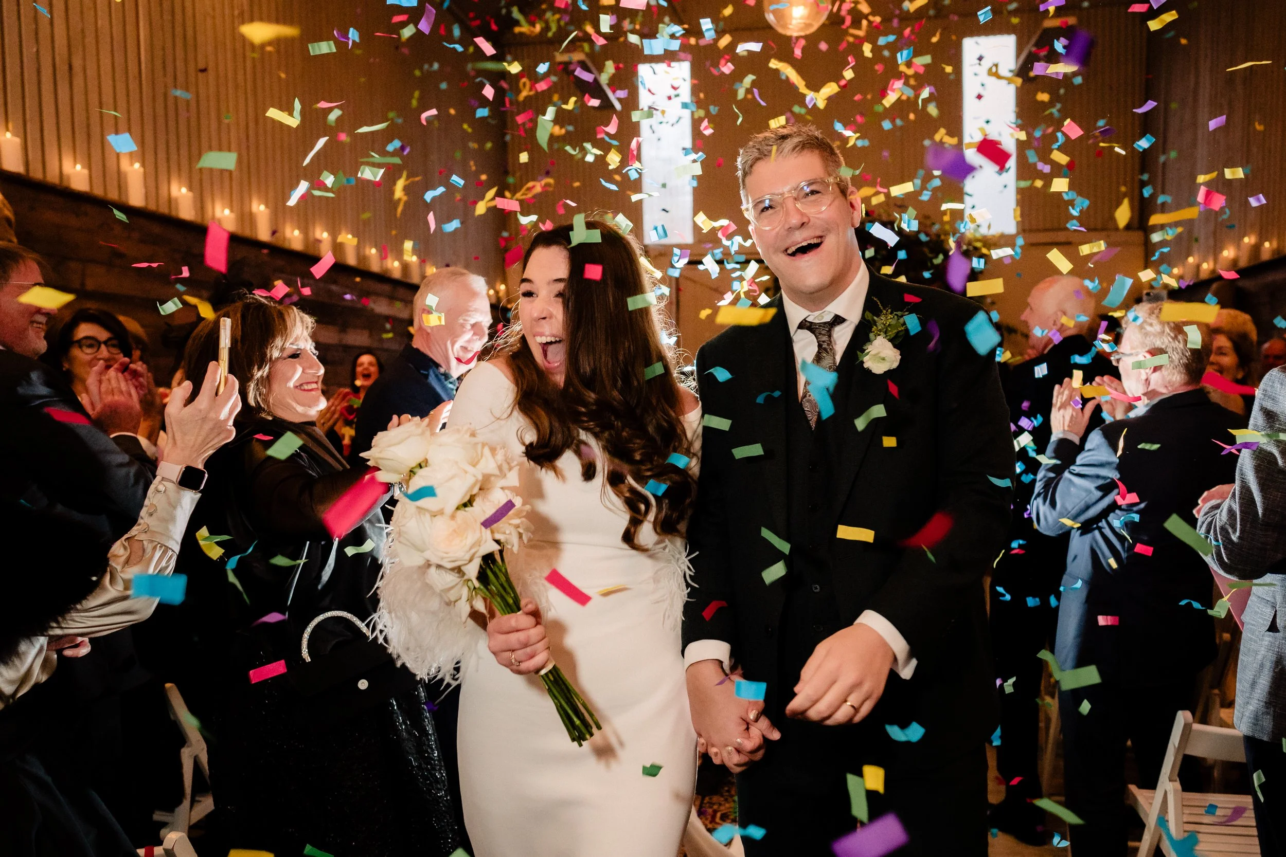 A wedding celebration with a bride and groom holding hands and smiling as colorful confetti falls around them, surrounded by seated guests applauding and celebrating inside a decorated venue.