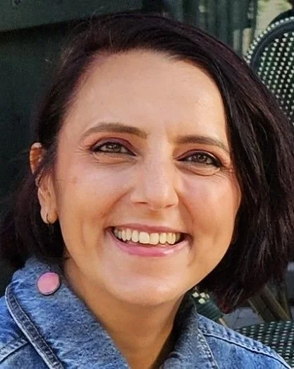 A woman with short dark hair smiling outdoors, wearing a denim jacket.