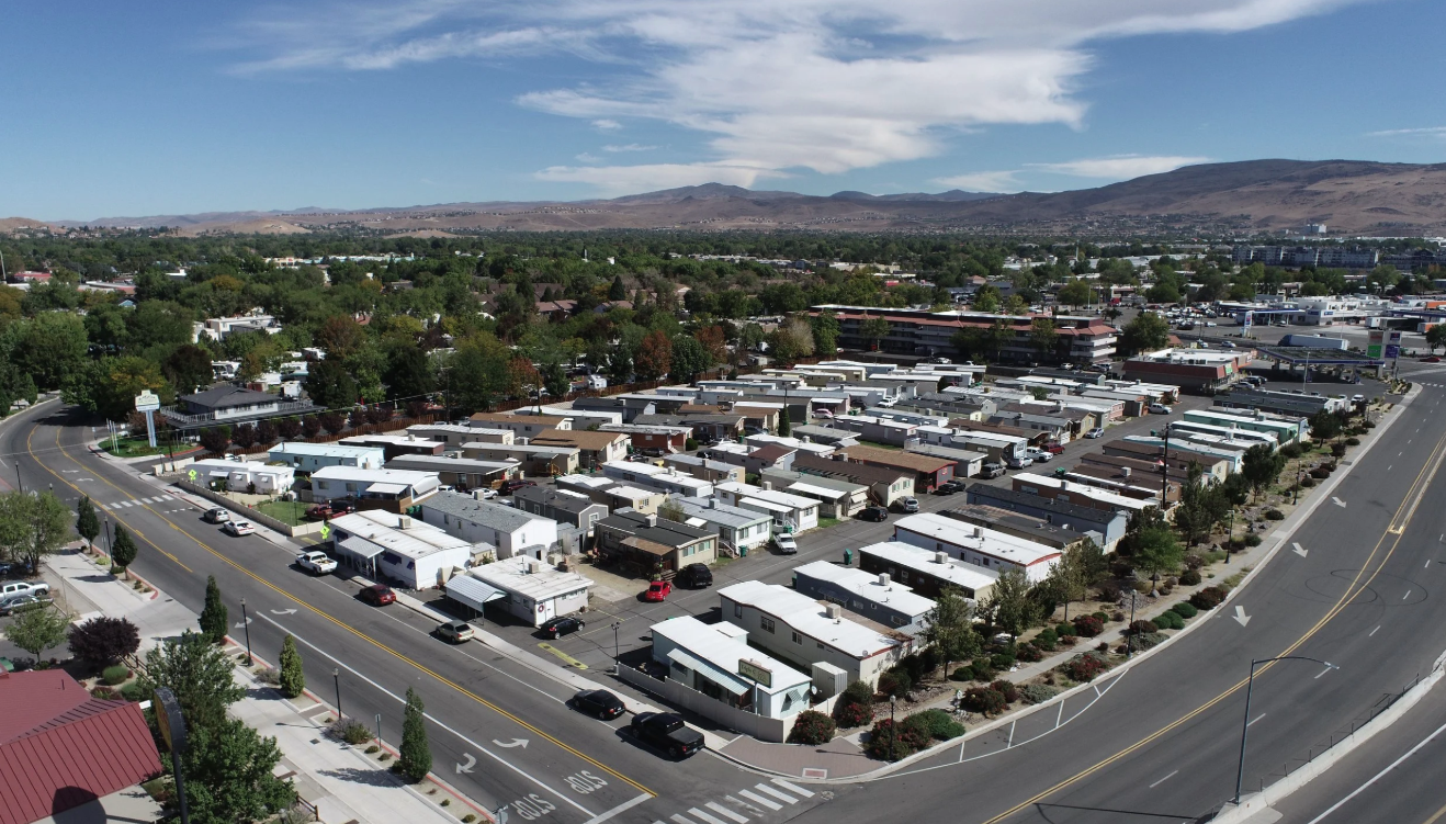 Aerial view of a residential mobile home park with multiple rows of mobile homes, surrounded by roads, trees, and a suburban area with mountains in the background.