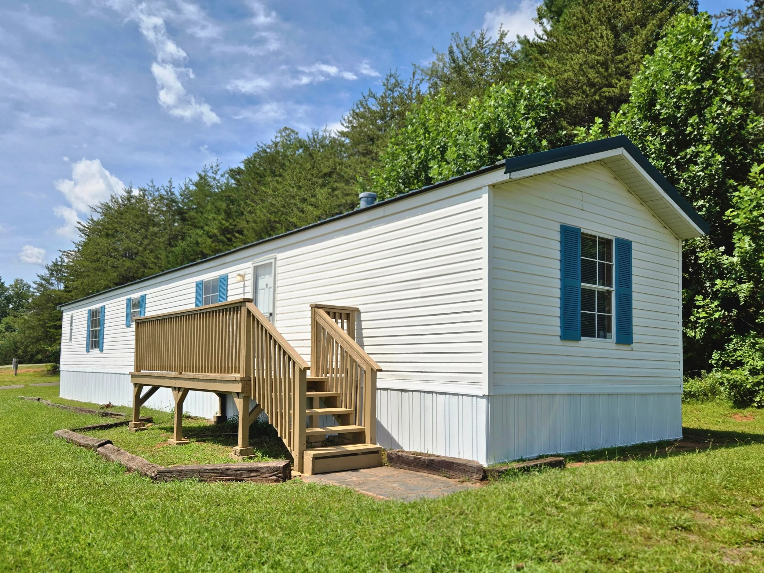 A white manufactured home with blue shutters, a small wooden front porch with stairs, set in a grassy yard with trees in the background and a blue sky with clouds overhead.