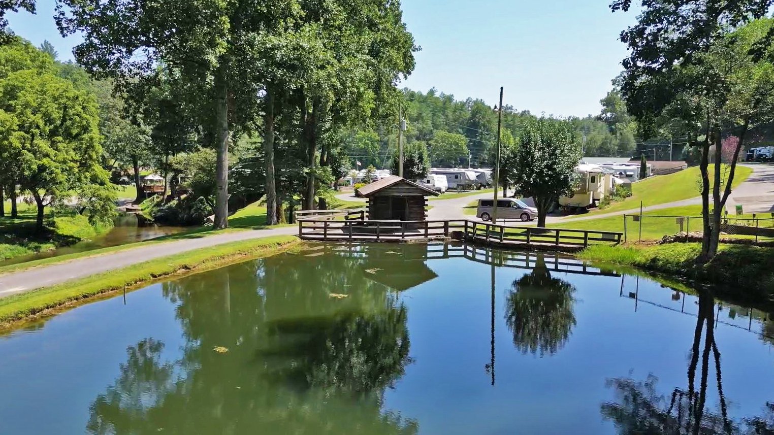 A small pond reflecting nearby trees and part of a wooden bridge near a small building, with lush green grass, trees, and recreational vehicles in the background on a sunny day.