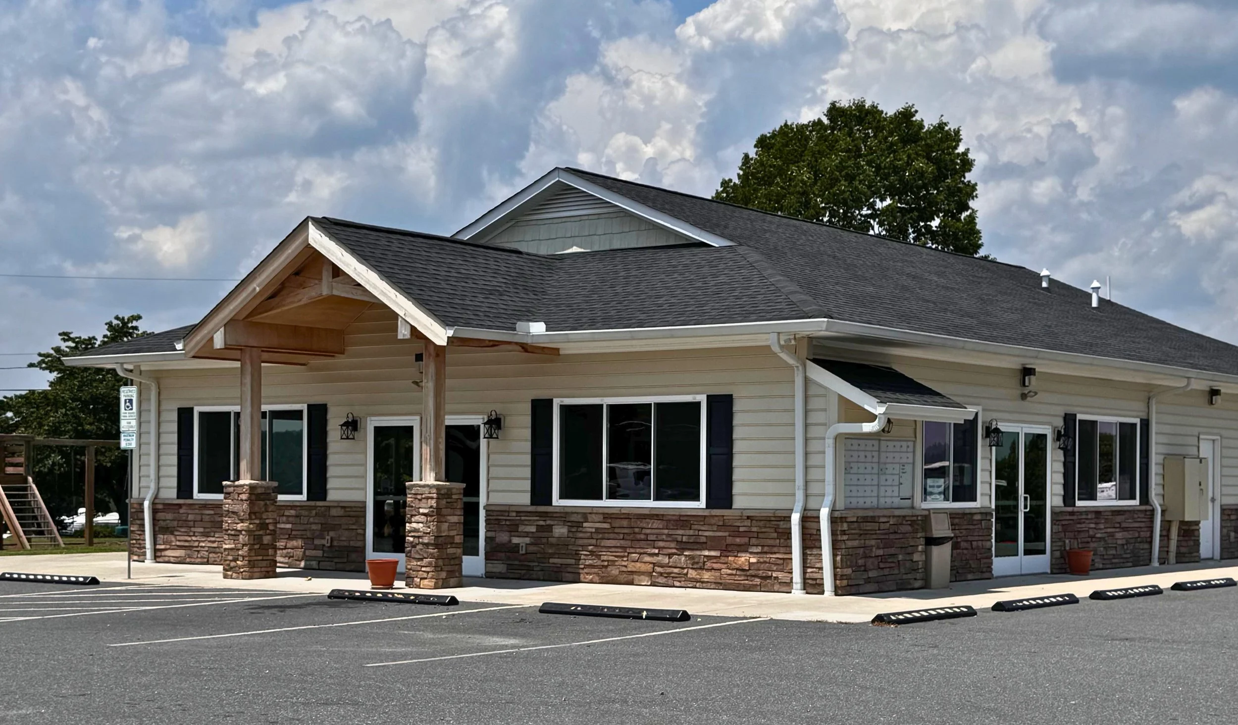 A small commercial building with a gray shingled roof, white siding, and a stone facade at the bottom. It has large windows with black shutters, a glass door, and a covered entrance supported by brick columns. The building is next to a parking lot wi