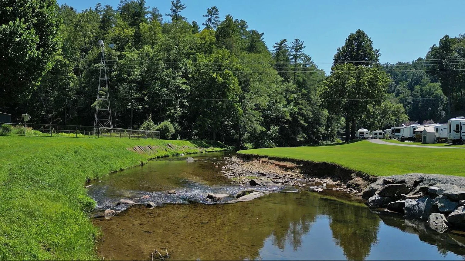 A small creek with clear water flowing through a grassy park area, surrounded by trees, with RVs parked on the right side and a blue sky overhead.