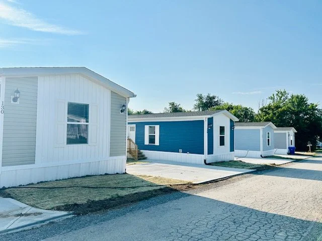 Row of new manufactured homes with different exterior colors along a paved street, under a clear blue sky.