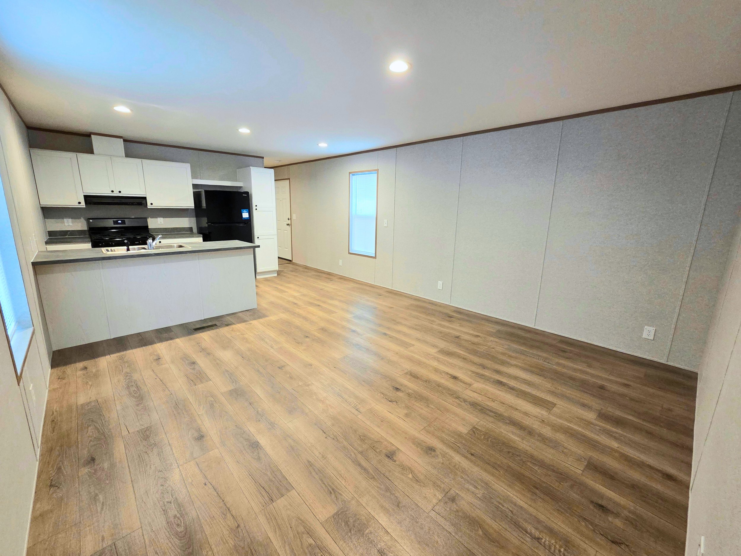 Empty living space with hardwood floors, kitchen area with white cabinets, black appliances, and a window on the wall.