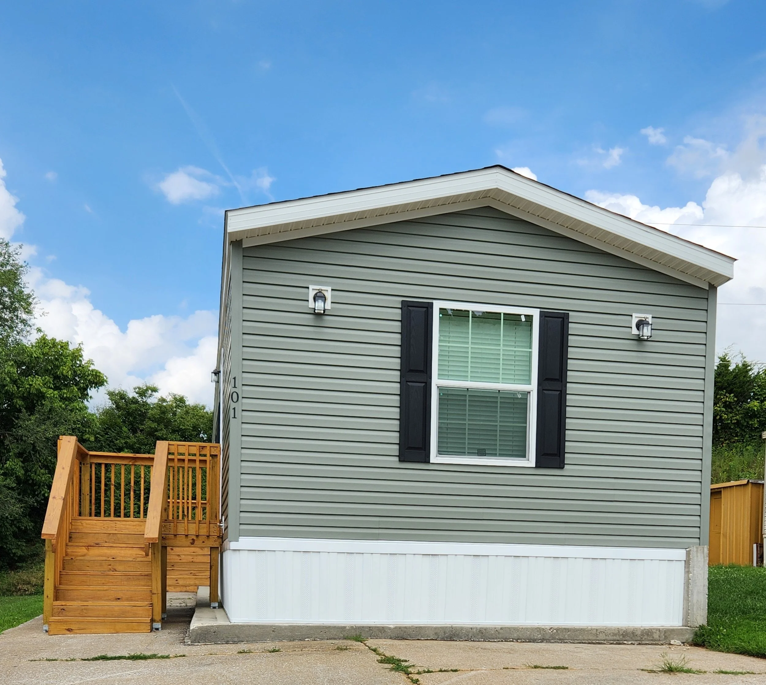 Front view of a modern house with gray siding, a single window with white blinds, black shutters, and a wooden staircase leading to the entrance. Bright blue sky with scattered clouds in the background.