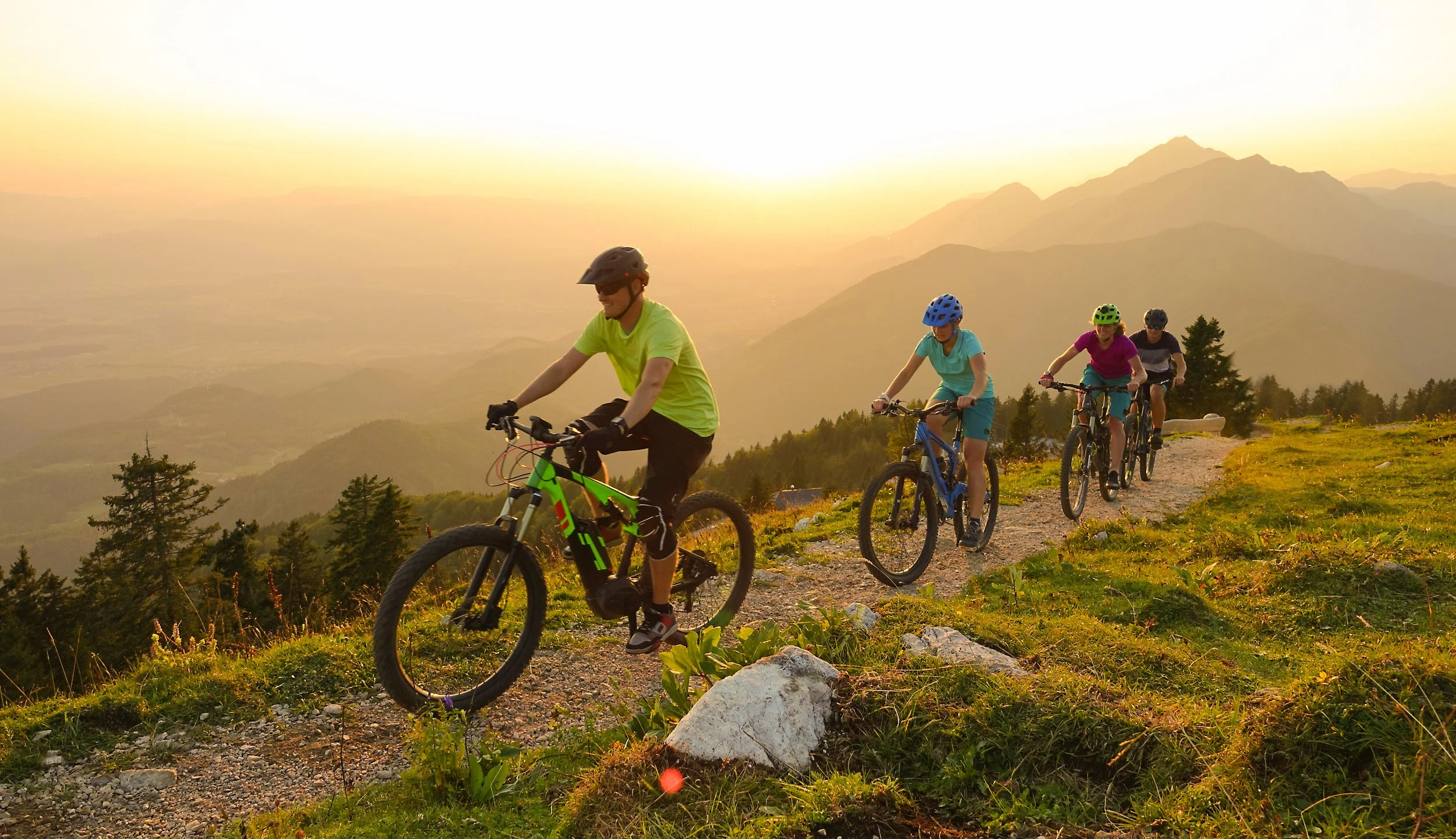 Group of four cyclists riding mountain bikes along a trail at sunset with mountains in the background