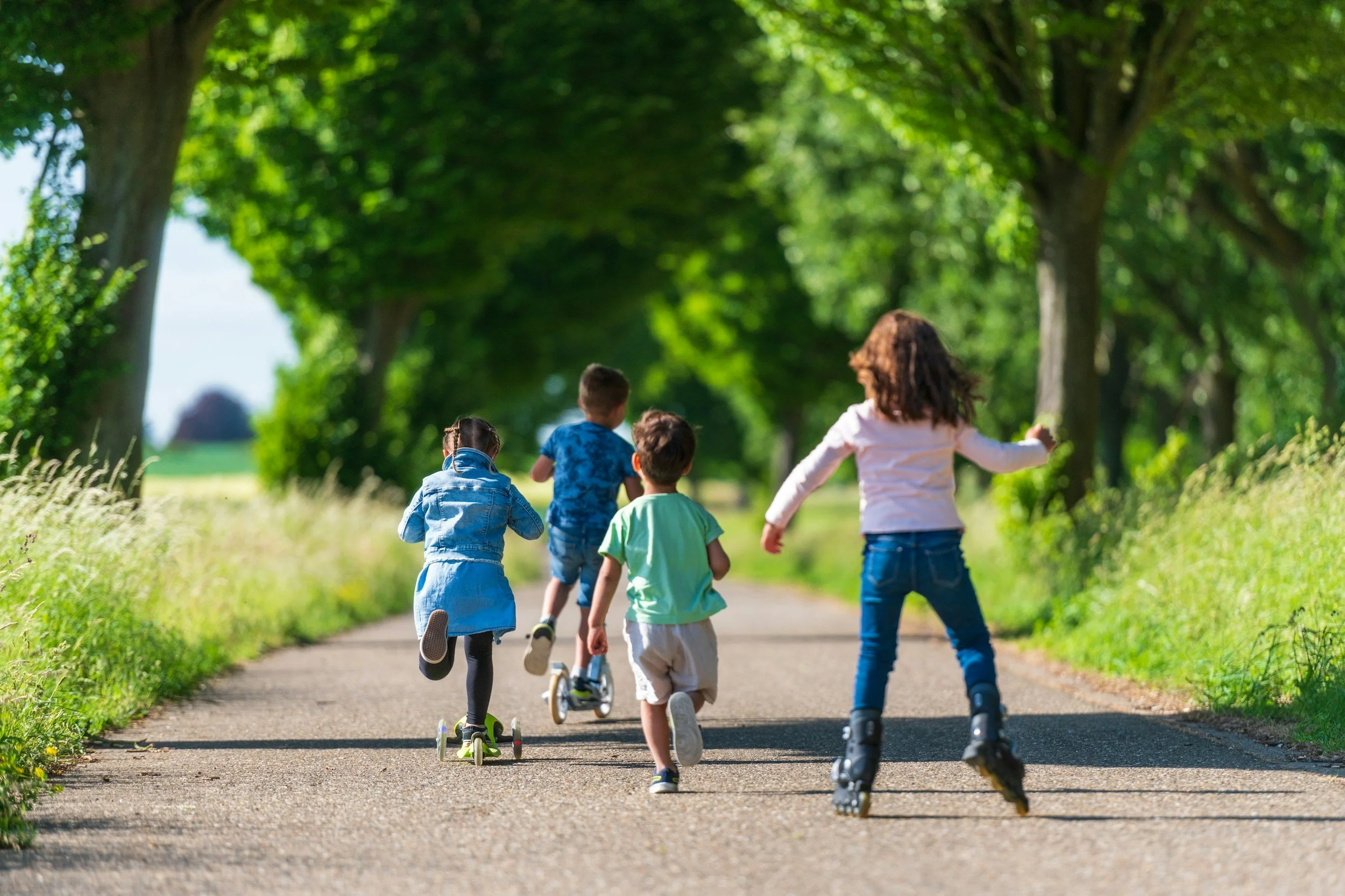 Group of children running and roller skating on a paved path in a green, tree-lined park on a sunny day.