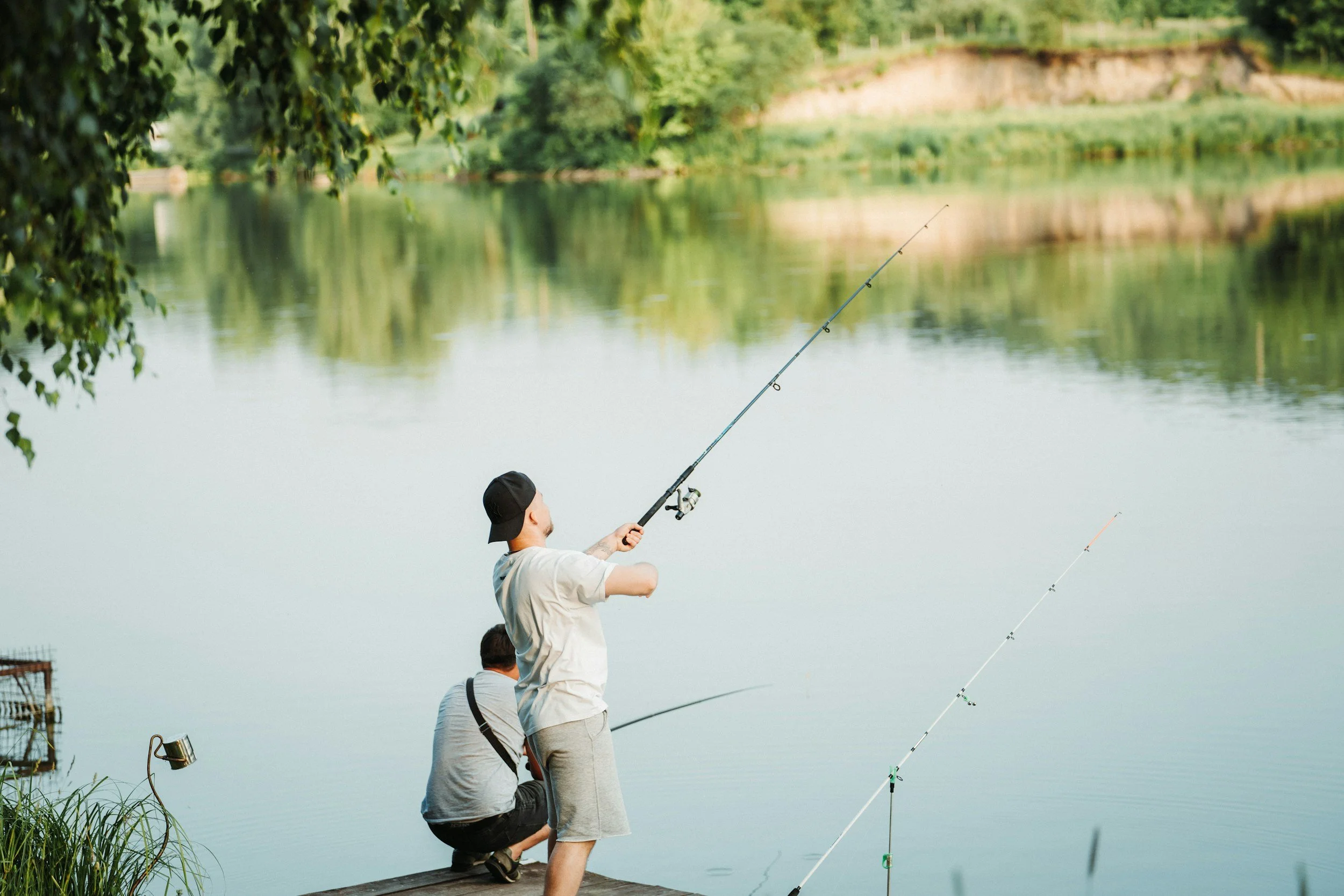 A boy and a man fishing by a lake on a wooden dock, with fishing rods cast into the water, surrounded by greenery and trees.