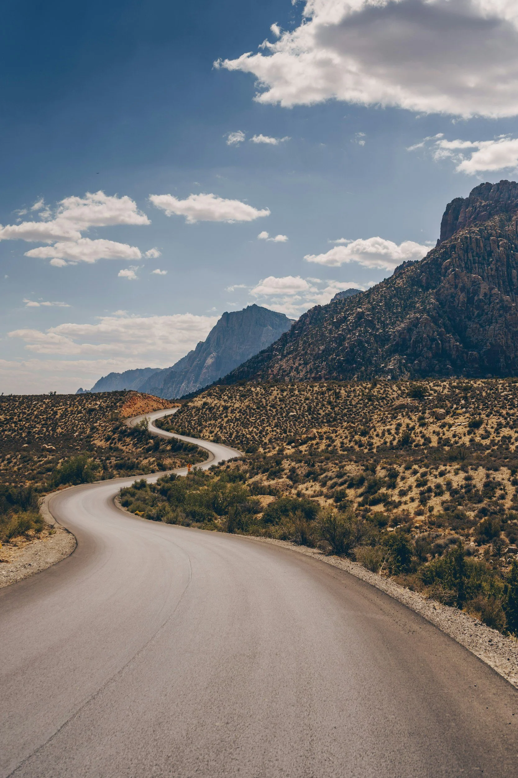A winding road through a desert landscape with mountains in the background and a partly cloudy sky overhead.