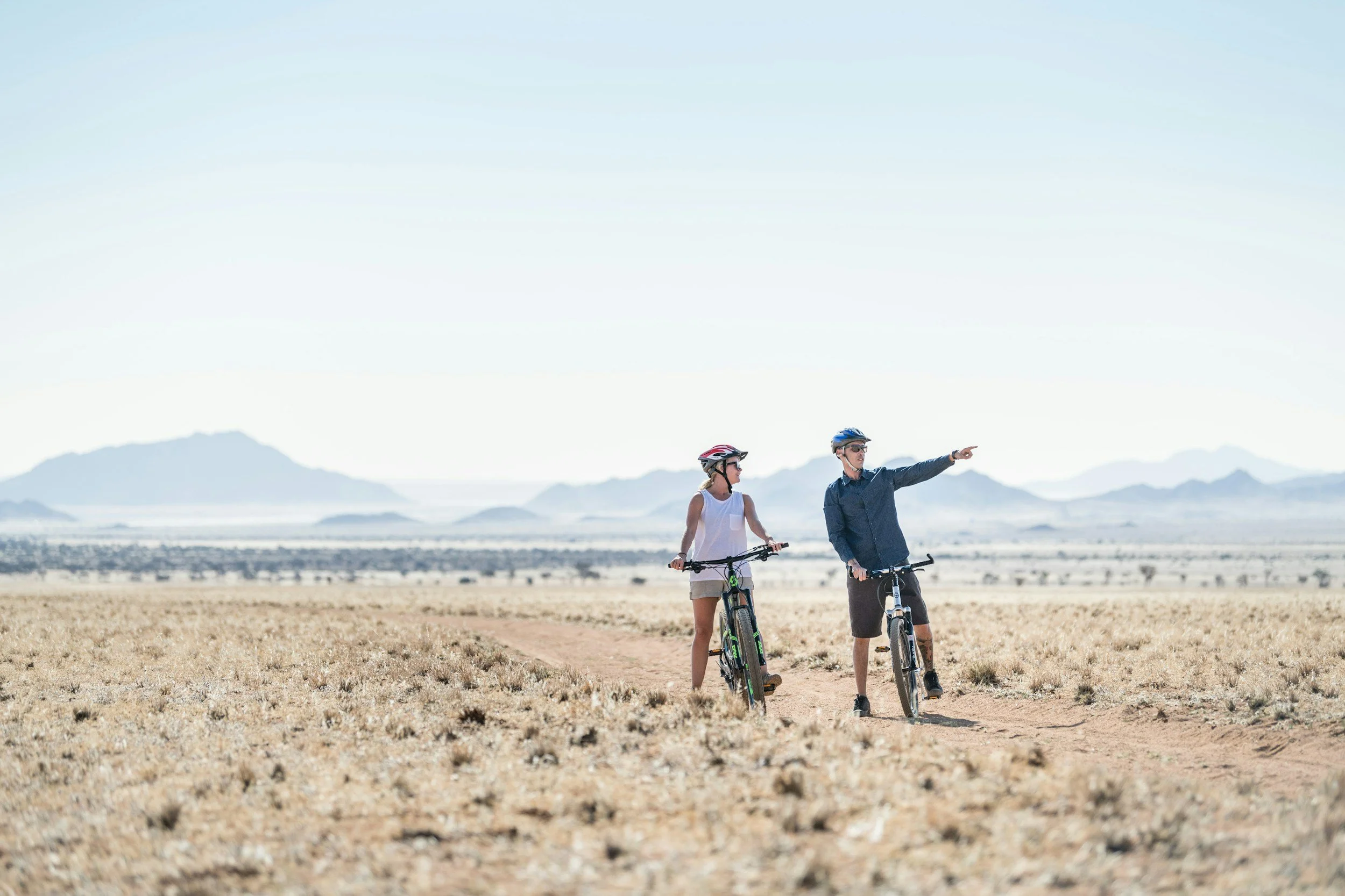 Two people in helmets standing with bicycles on a dirt trail in a desert landscape, mountains in the background, one person pointing ahead.