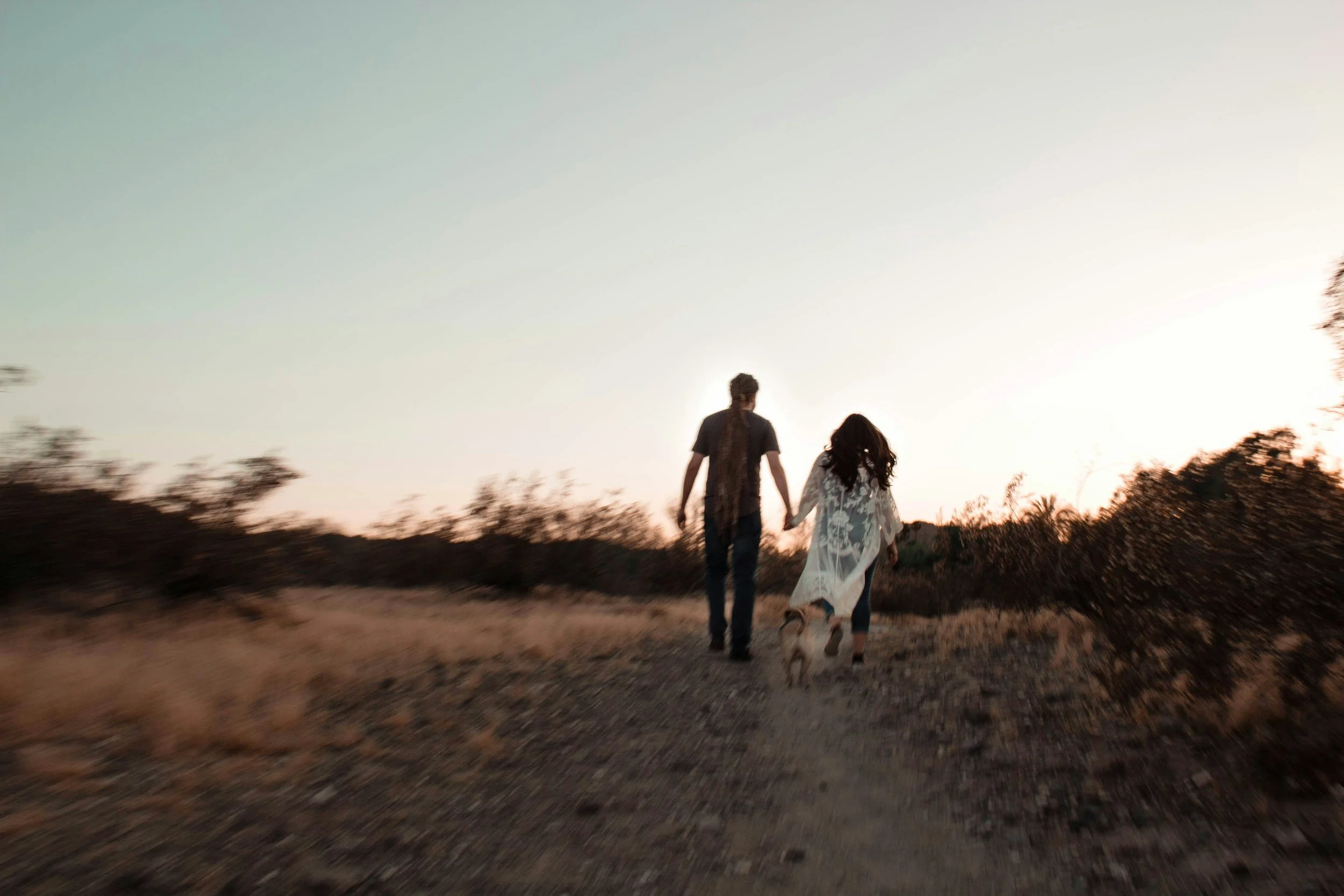 A couple walking hand in hand on a dirt path through a grassy field with bushes, at sunset or sunrise, with a small dog walking nearby.