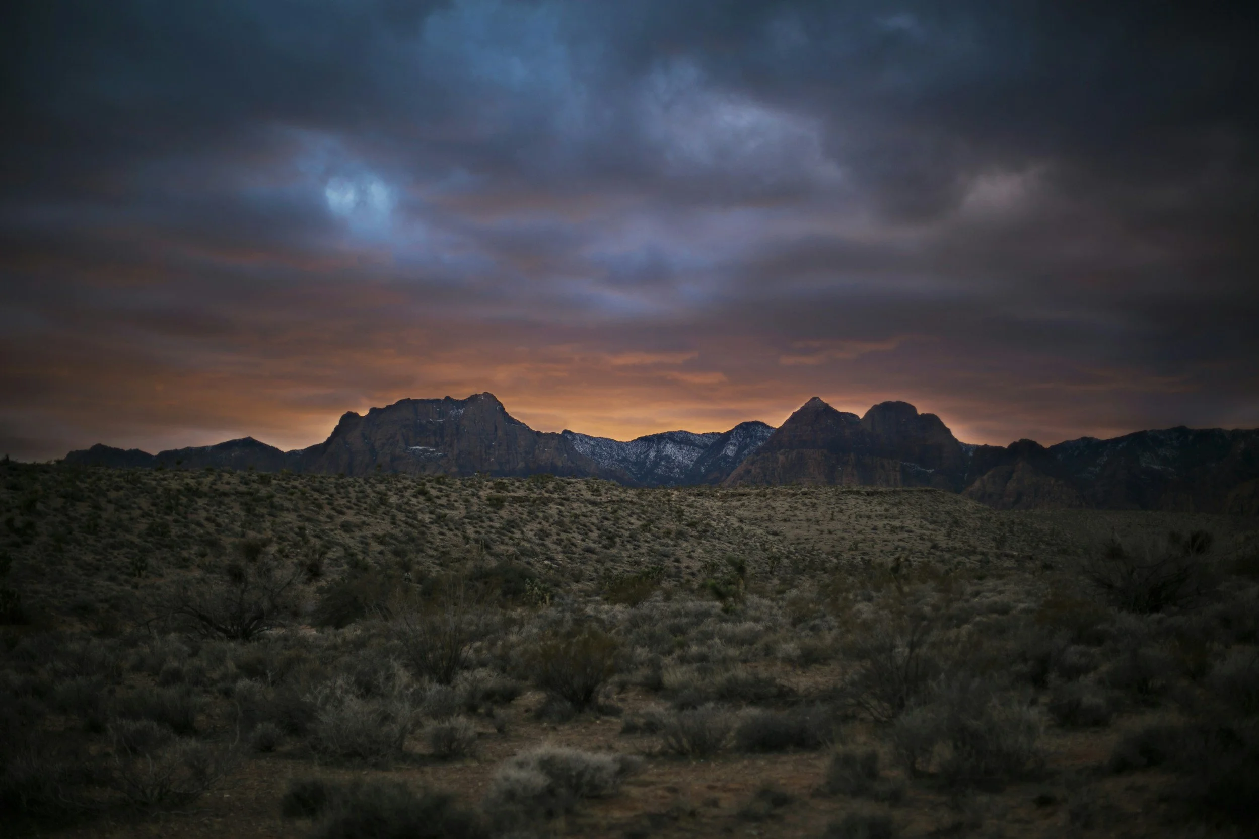 Desert landscape at sunset with rugged mountains and layered dark clouds in the sky.