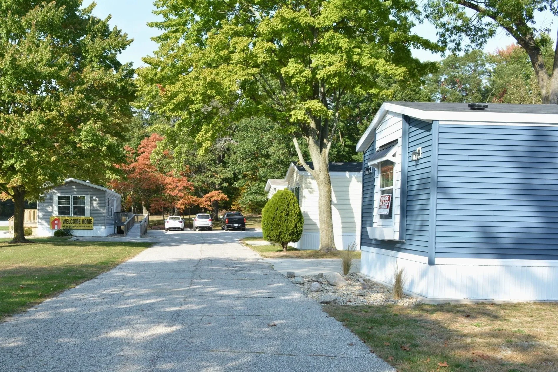 Residential mobile home park with several houses, trees, and parked cars, with a paved road in the foreground.