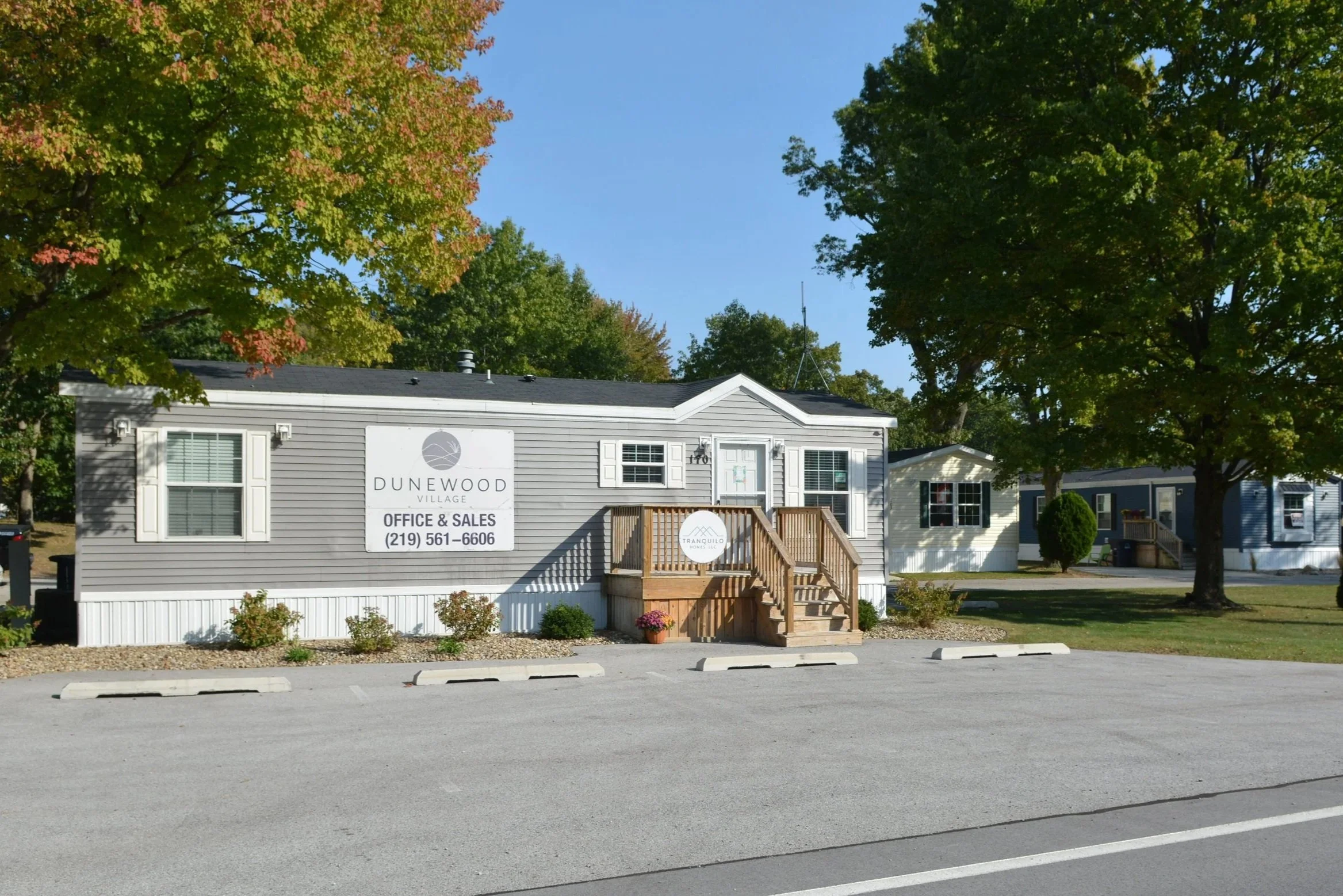 Single-story gray building with white trim and a small wooden porch, surrounded by trees with green and some autumn-colored leaves, in a parking lot with marked spaces.