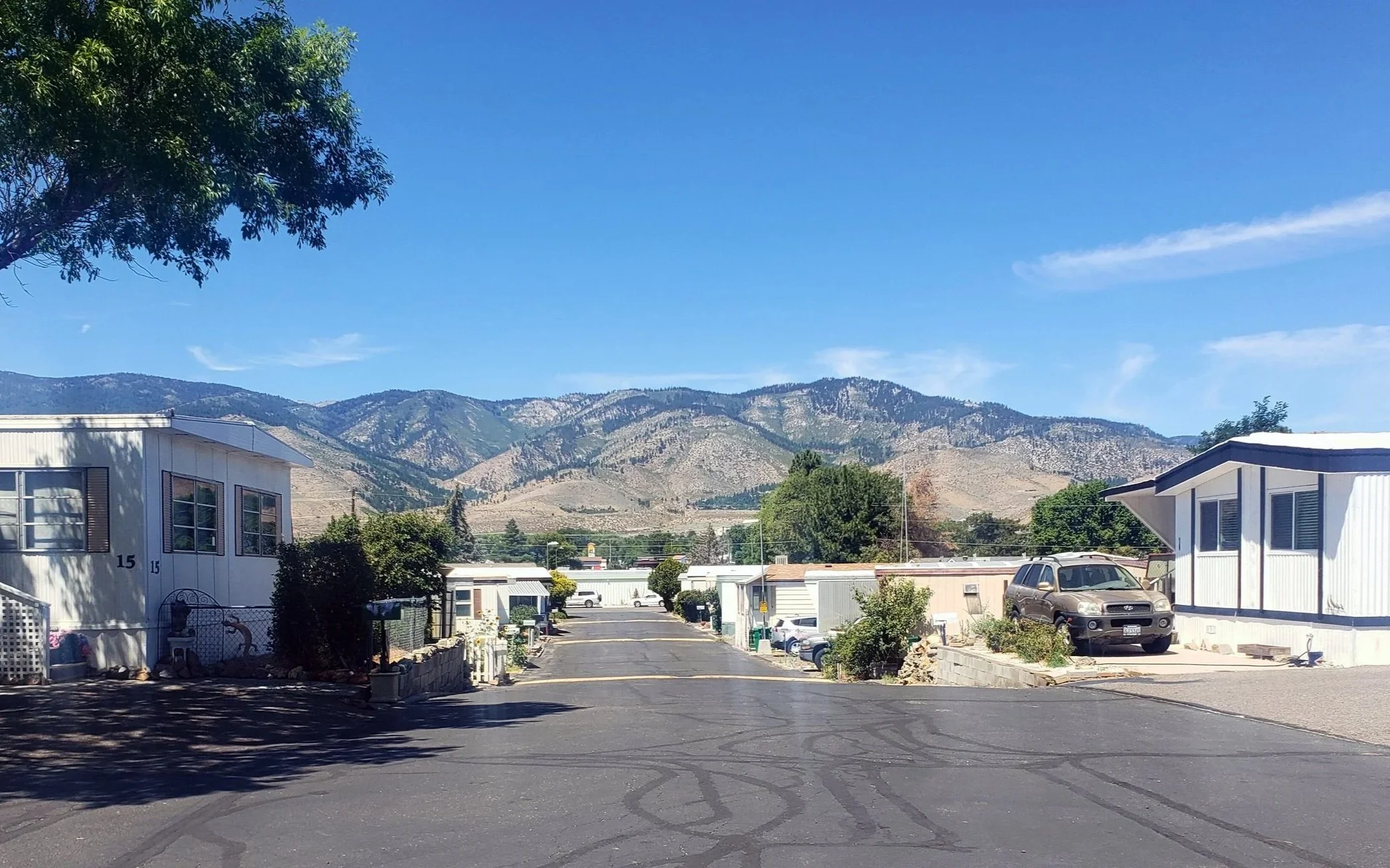 Mobile home park with mobile homes, parked cars, trees, and mountainous landscape in the background under a clear blue sky.
