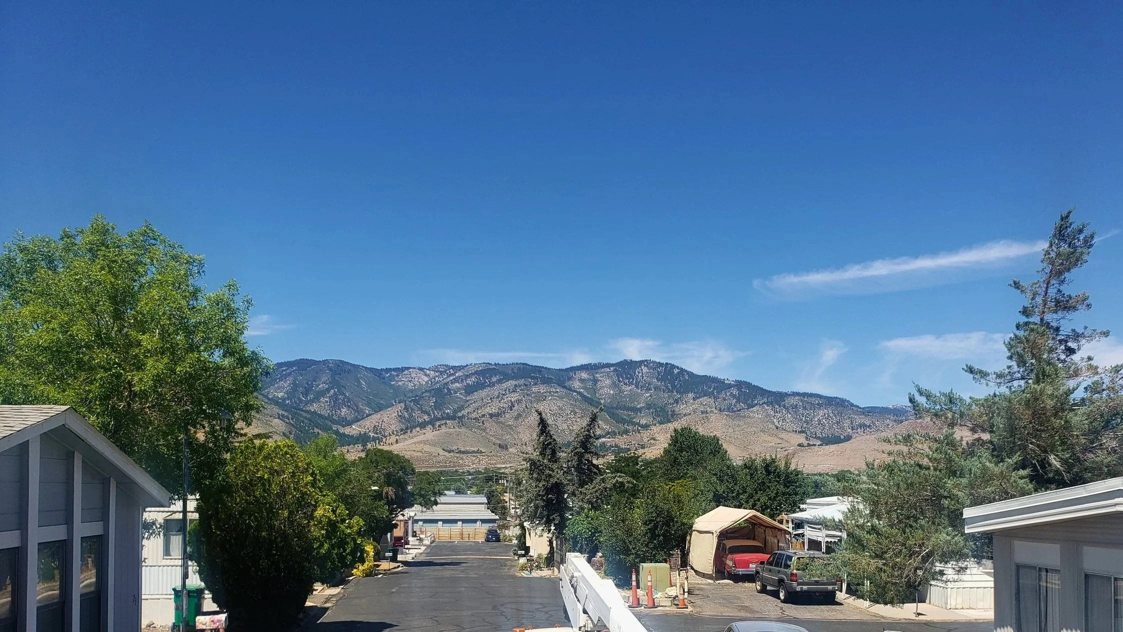 View of a parking lot with utility trucks and various cars, surrounded by mobile homes, trees, and mountains in the background under a blue sky.
