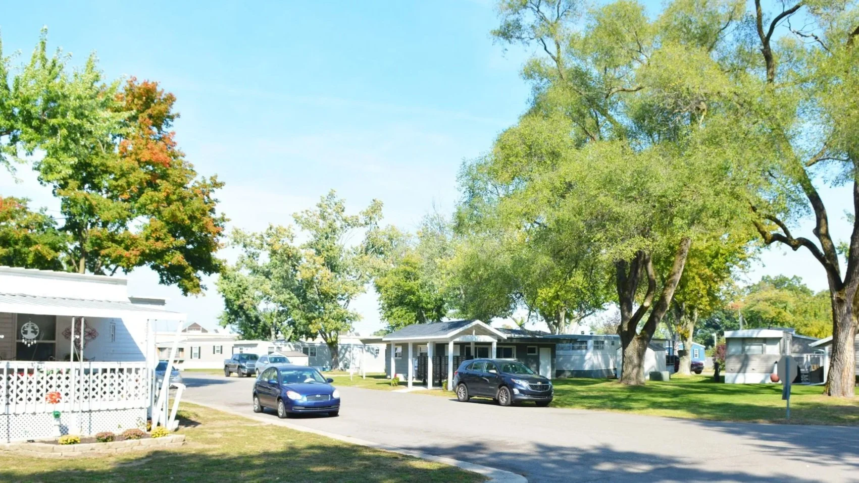 A peaceful mobile home park with rows of mobile homes, a paved road, and parked cars under large green trees on a sunny day.