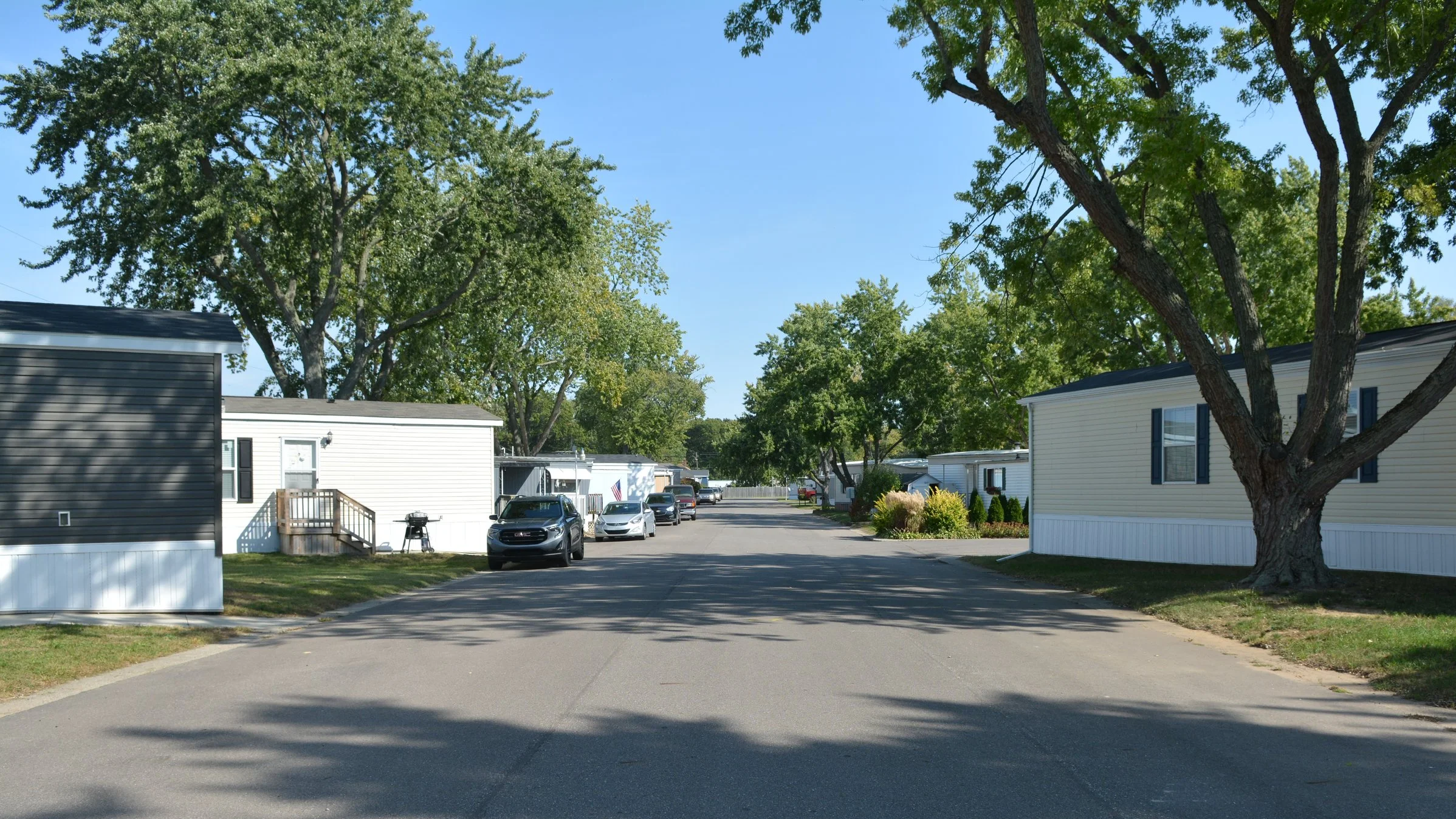 A residential neighborhood street with mobile homes on both sides, parked cars, and large trees providing shade on a sunny day.