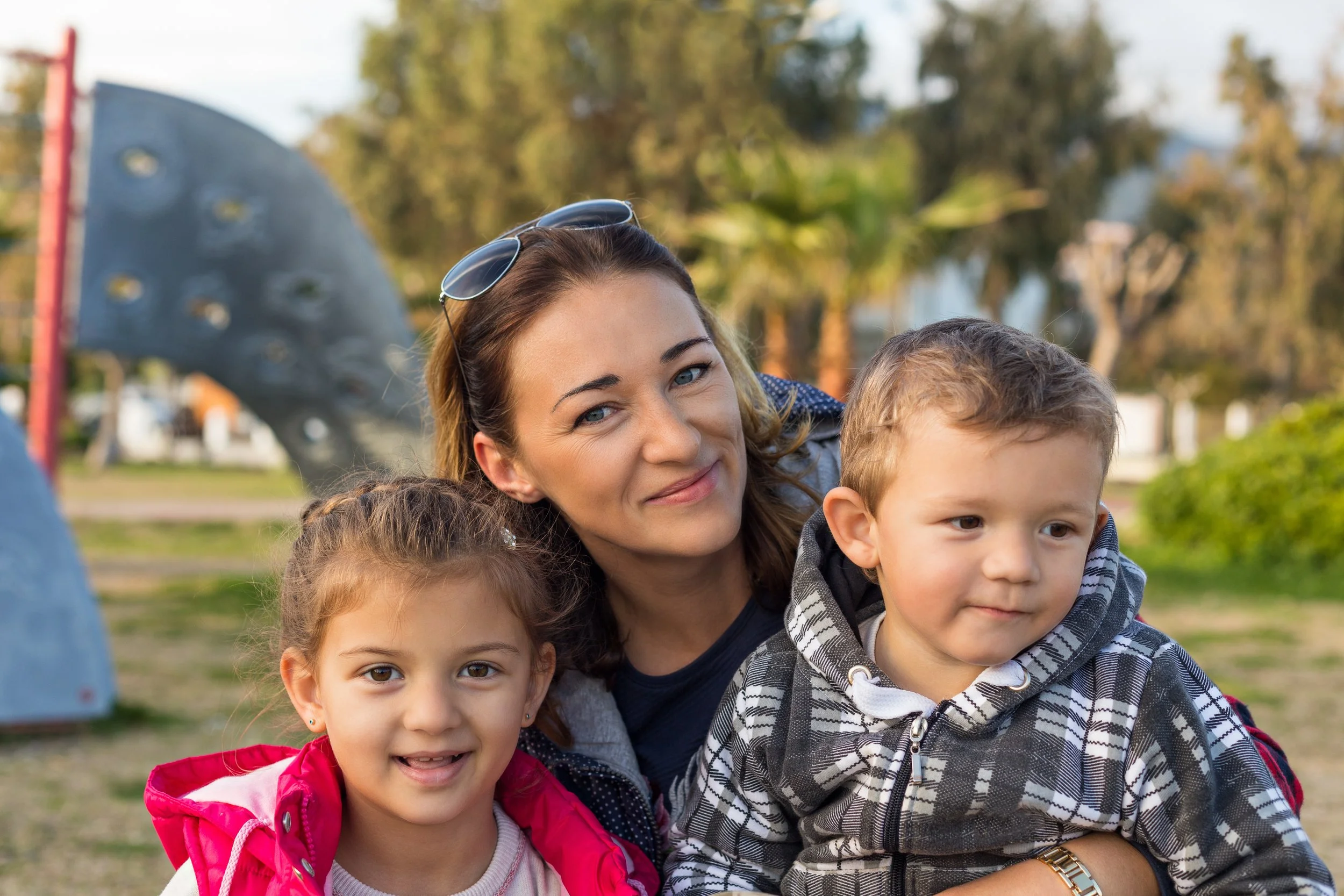 A woman with sunglasses on her head smiling with a young girl and boy outdoors in a park on a sunny day.