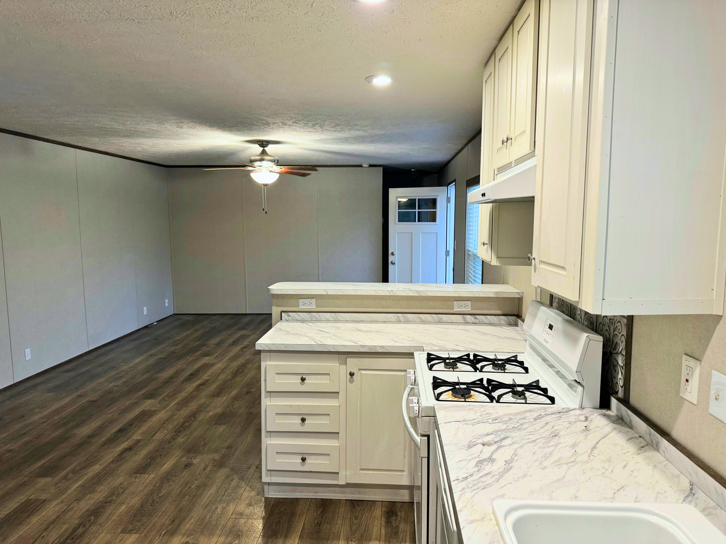 Empty living room and kitchen with white cabinets, marble countertops, wood flooring, ceiling fan, and front door.