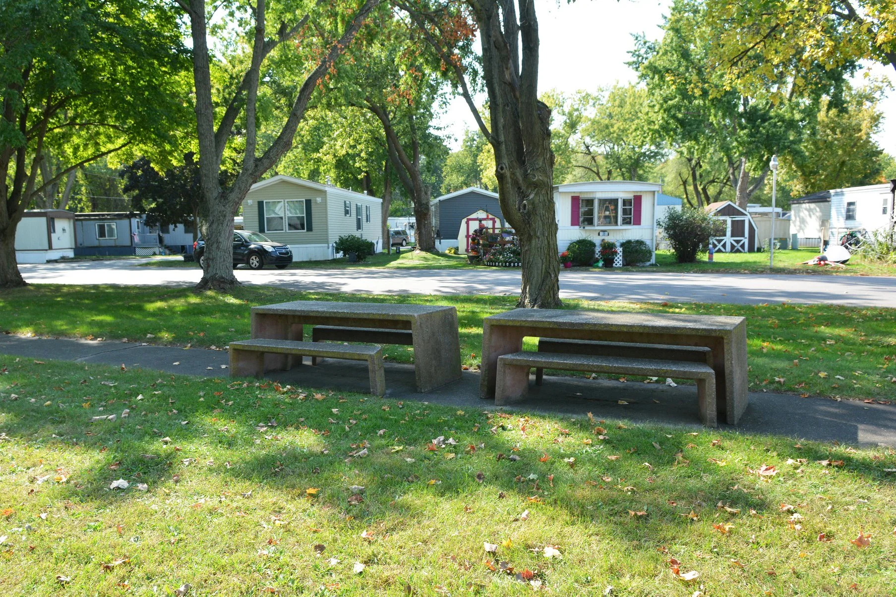 A park with concrete benches and trees, with a row of mobile homes and a car parked on the street in the background.