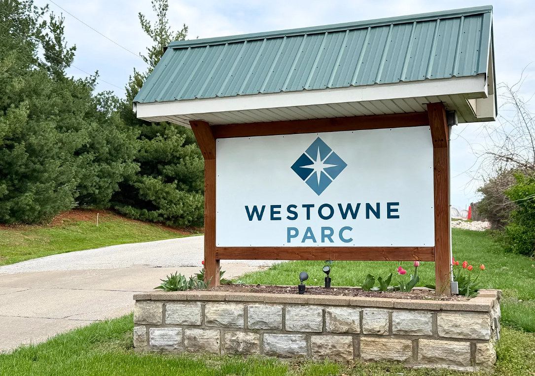 Sign for Westowne Park with a blue and white logo, situated on a stone border, with a driveway and green trees in the background.