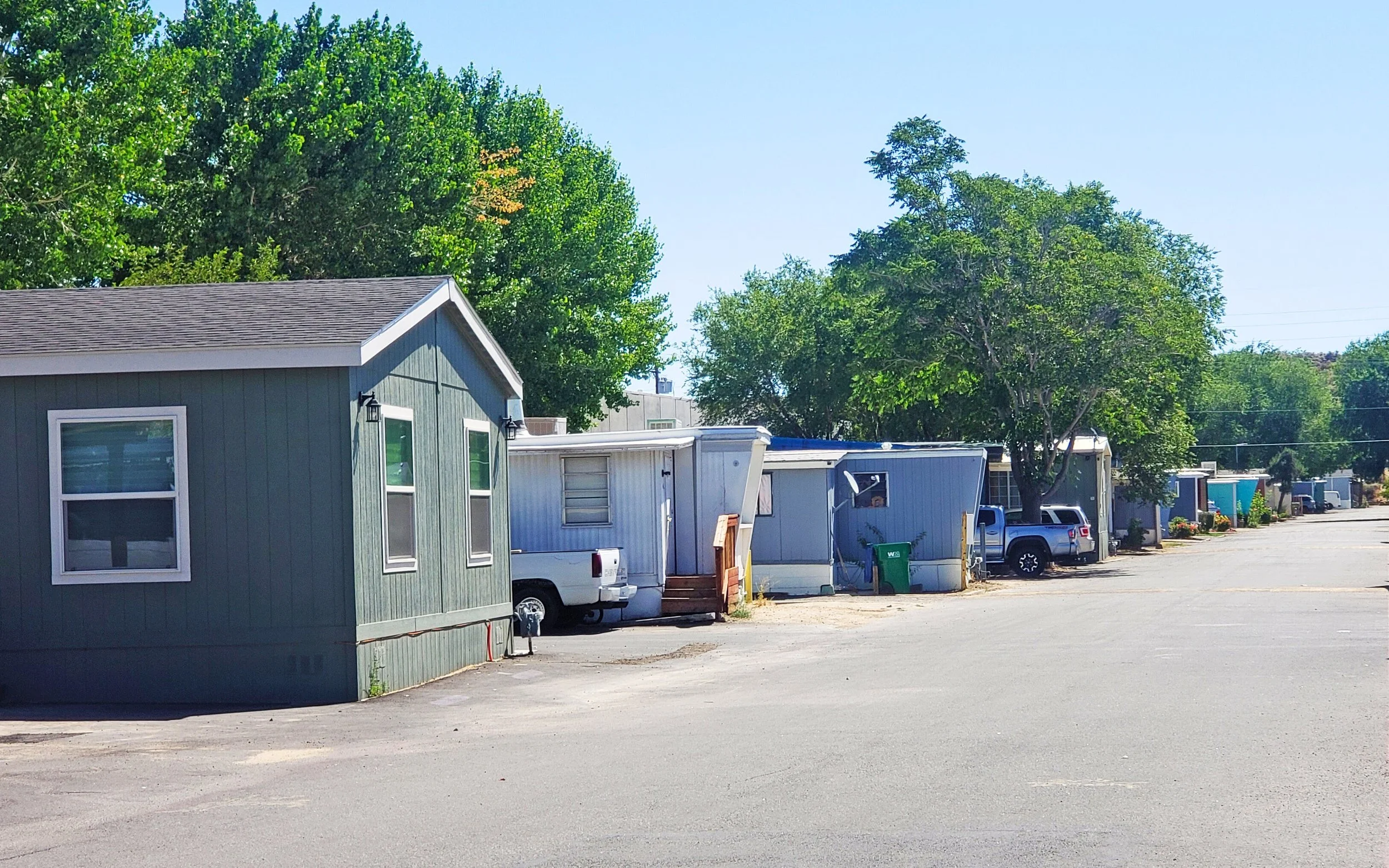 A row of mobile homes and RVs parked along a wide street with trees and a clear blue sky in the background.
