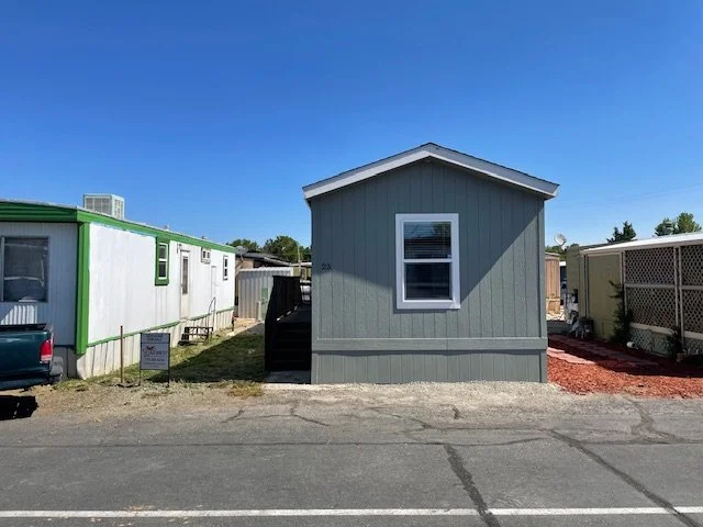 Small gray house with a white-framed window on a paved lot, with mobile homes and trailers on either side under a clear blue sky.