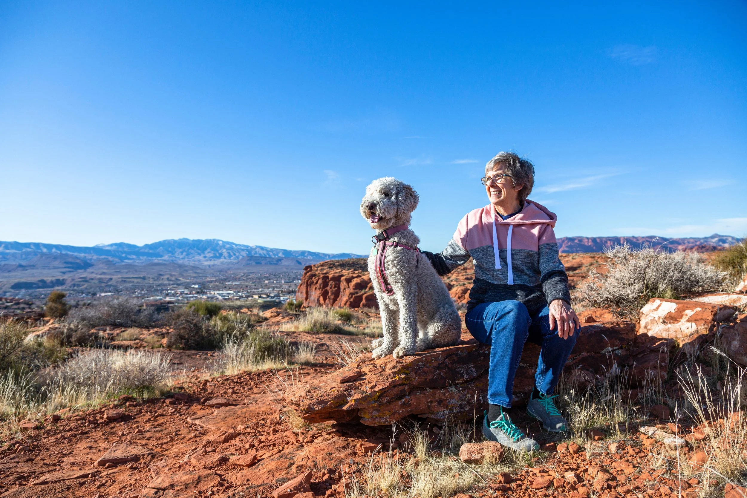 A woman with gray hair, glasses, and a pink, gray, and black hoodie sitting on a red rocky hill with a large white poodle, with a desert landscape and mountains in the background.