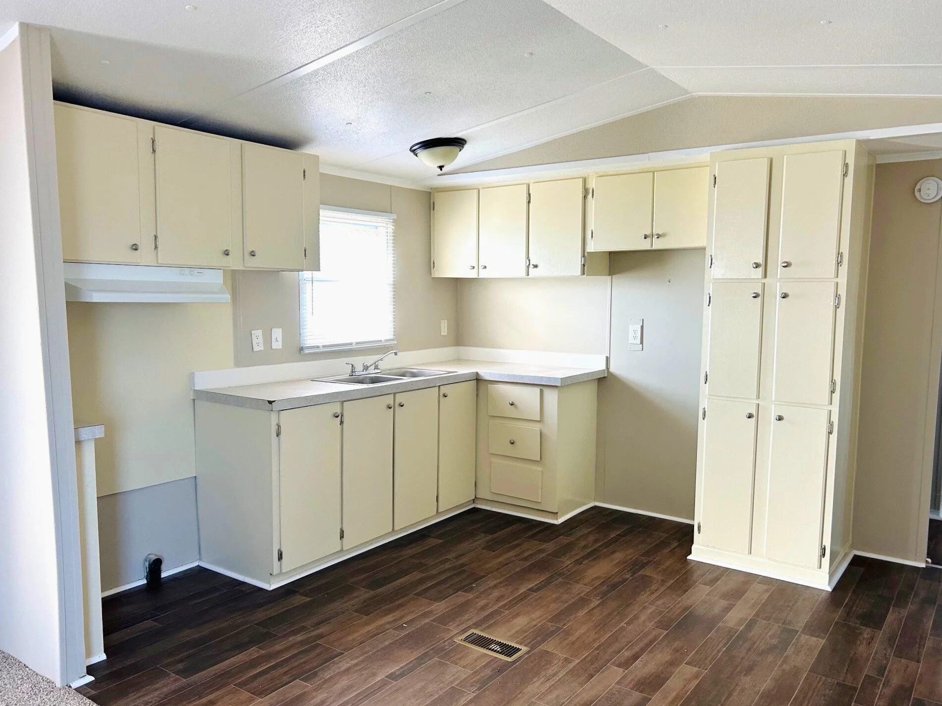 Empty kitchen with cream cabinets, white countertops, a window with blinds, and dark wood flooring.