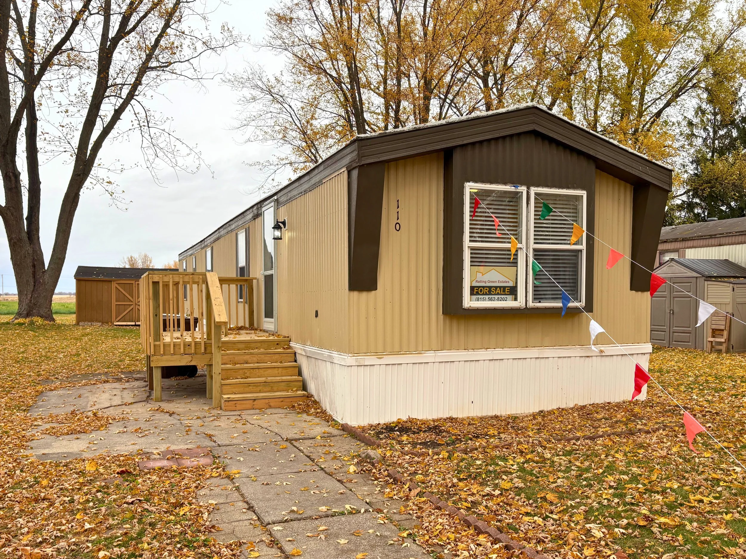 A manufactured home with a small wooden porch and stairs, for sale sign in the window, surrounded by fallen autumn leaves, with a large tree and open field in the background.