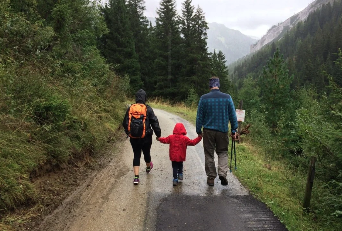 A family of three, including two adults and one child, walks on a dirt and paved trail in a forested mountainous area during misty weather. The child is holding both adult's hands.